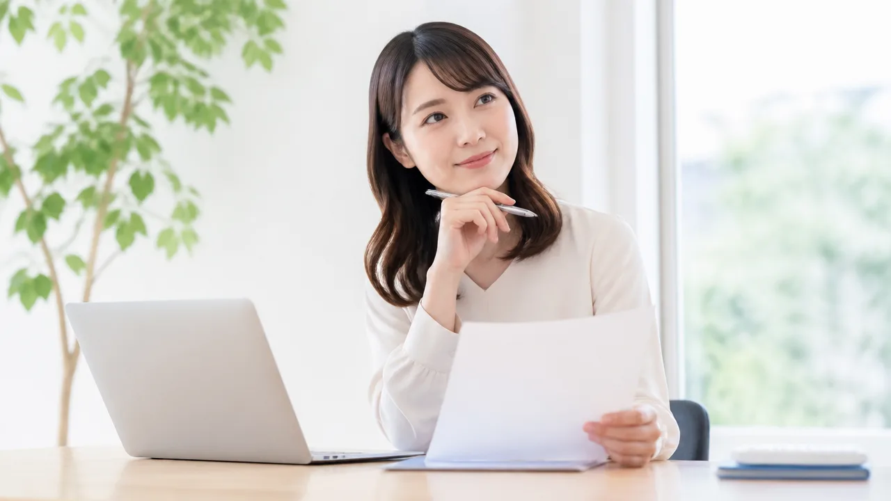 ノートパソコンの前で書類を手に、内容を考える人のイメージ