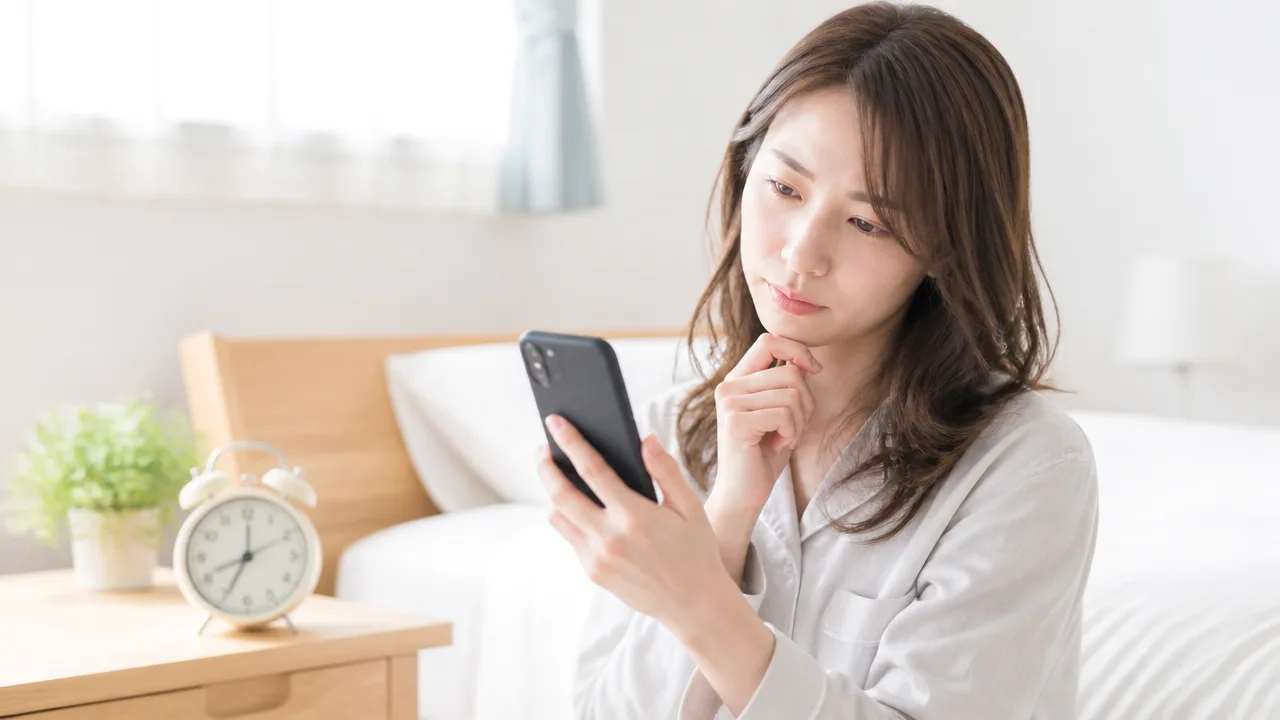 ベッドのそばで、目覚まし時計とスマホを確認する人